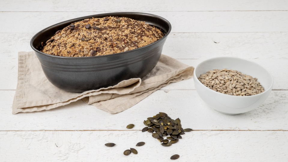 Körnerbrot in einer ovalen Brotbackform, daneben einige Kürbiskerne und ein Schälchen mit Sonnenblumenkernen Körnerbrot in einer ovalen Brotbackform, daneben einige Kürbiskerne und ein Schälchen mit Sonnenblumenkernen