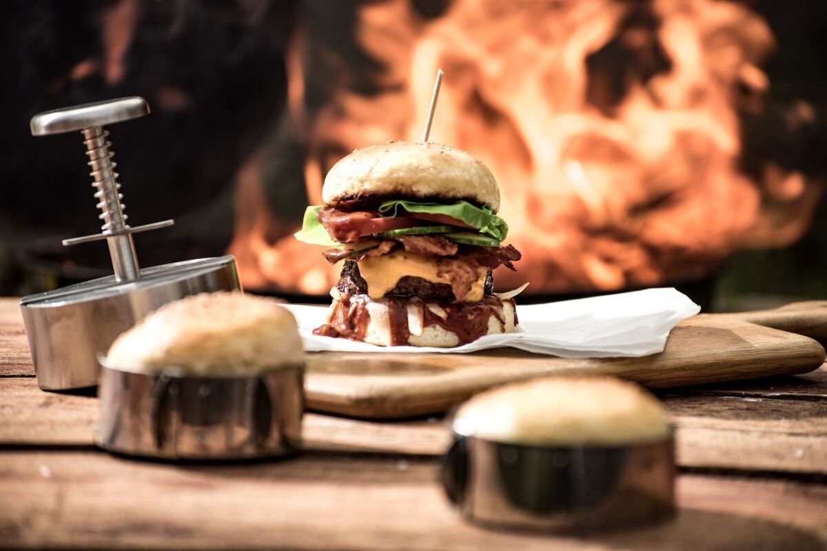 Burger Patty selber machen: Fertiger Burger, Burgerpresse und Buns vor flammendem Hintergrund Burger Patty selber machen: Fertiger Burger, Burgerpresse und Buns vor flammendem Hintergrund