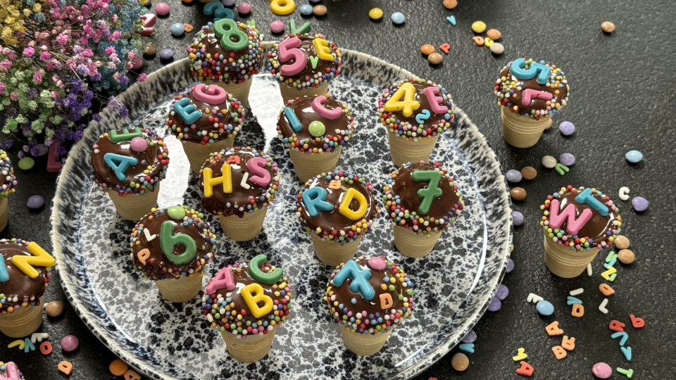 Kleine Kuchen mit Schokoladenglasur und Buchstaben-Dekoration im Waffelbecher auf einem Teller Kleine Kuchen mit Schokoladenglasur und Buchstaben-Dekoration im Waffelbecher auf einem Teller
