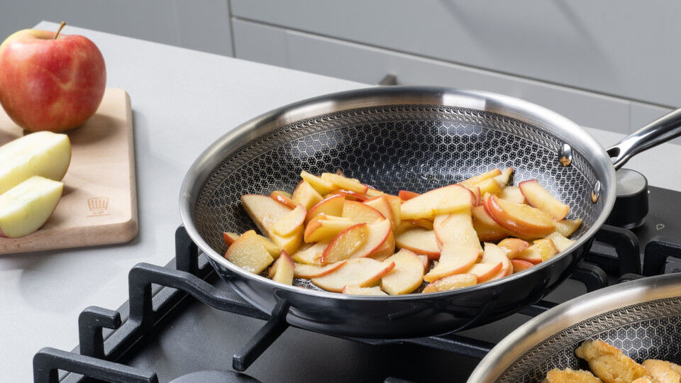 Apfelstücke in einer tiefen Pfanne auf dem Herd, daneben ein ganzer Apfel und ein aufgeschnittener Apfel auf einem Holz-Schneidebrett