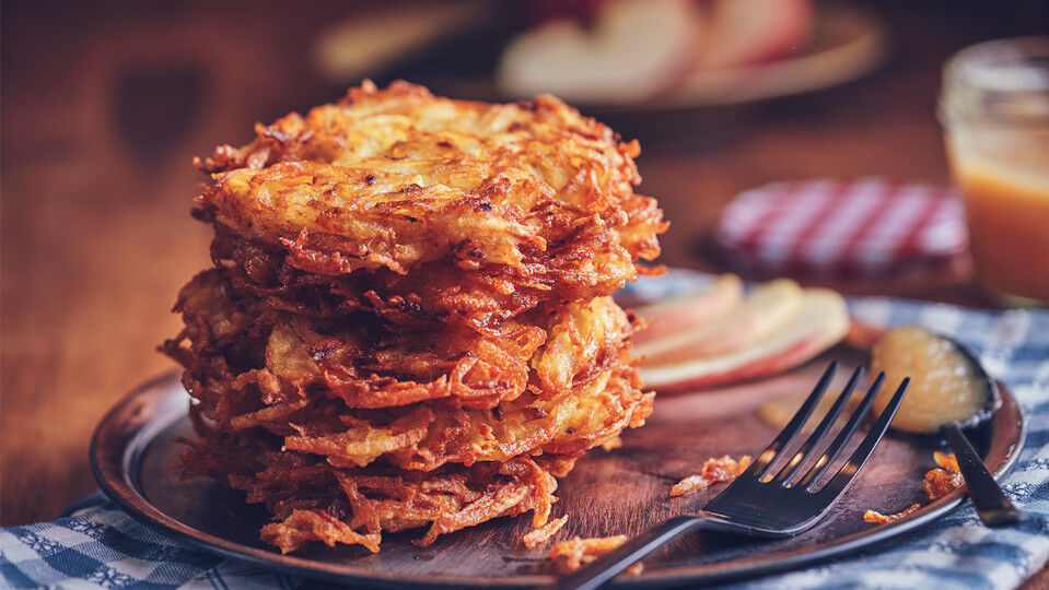 Ein Stapel aus mehreren Kartoffelpuffern auf einem Teller, daneben einige dünne Apfelscheiben und ein Löffel mit Apfelmus