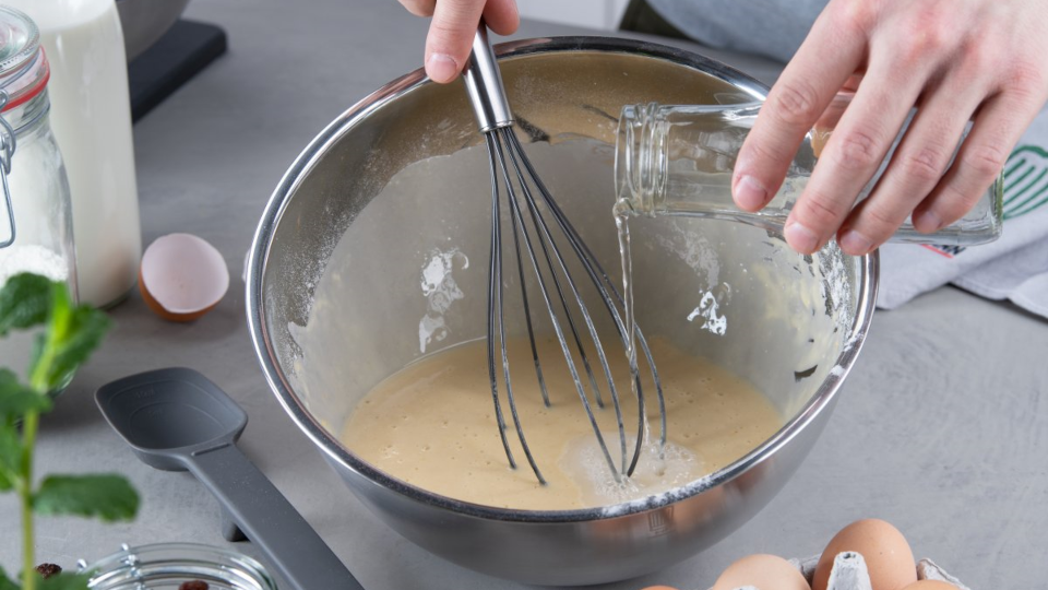 Rührschüssel mit Teig darin, unter den mit einem Schneebesen eine Flüssigkeit eingerührt wird Rührschüssel mit Teig darin, unter den mit einem Schneebesen eine Flüssigkeit eingerührt wird