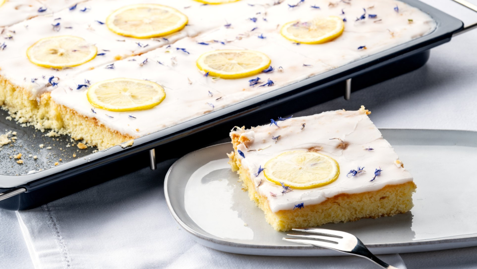 Zitronenkuchen mit Zuckerguss auf dem Backblech, daneben ein Stück auf einem Teller Zitronenkuchen mit Zuckerguss auf dem Backblech, daneben ein Stück auf einem Teller