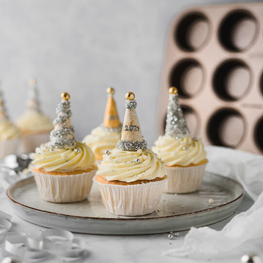 Silvester Cupcakes mit glitzernder Deko auf einer Tortenplatte, im Hintergrund ein Muffinblech