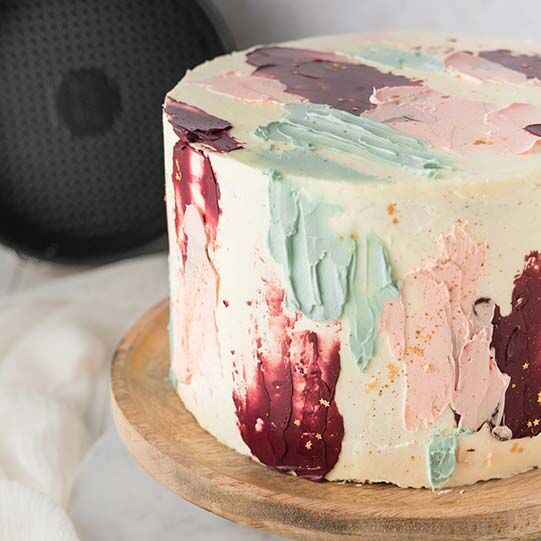 Bunte Flecken-Torte auf einer Tortenplatte aus Holz, im Hintergrund eine Springform