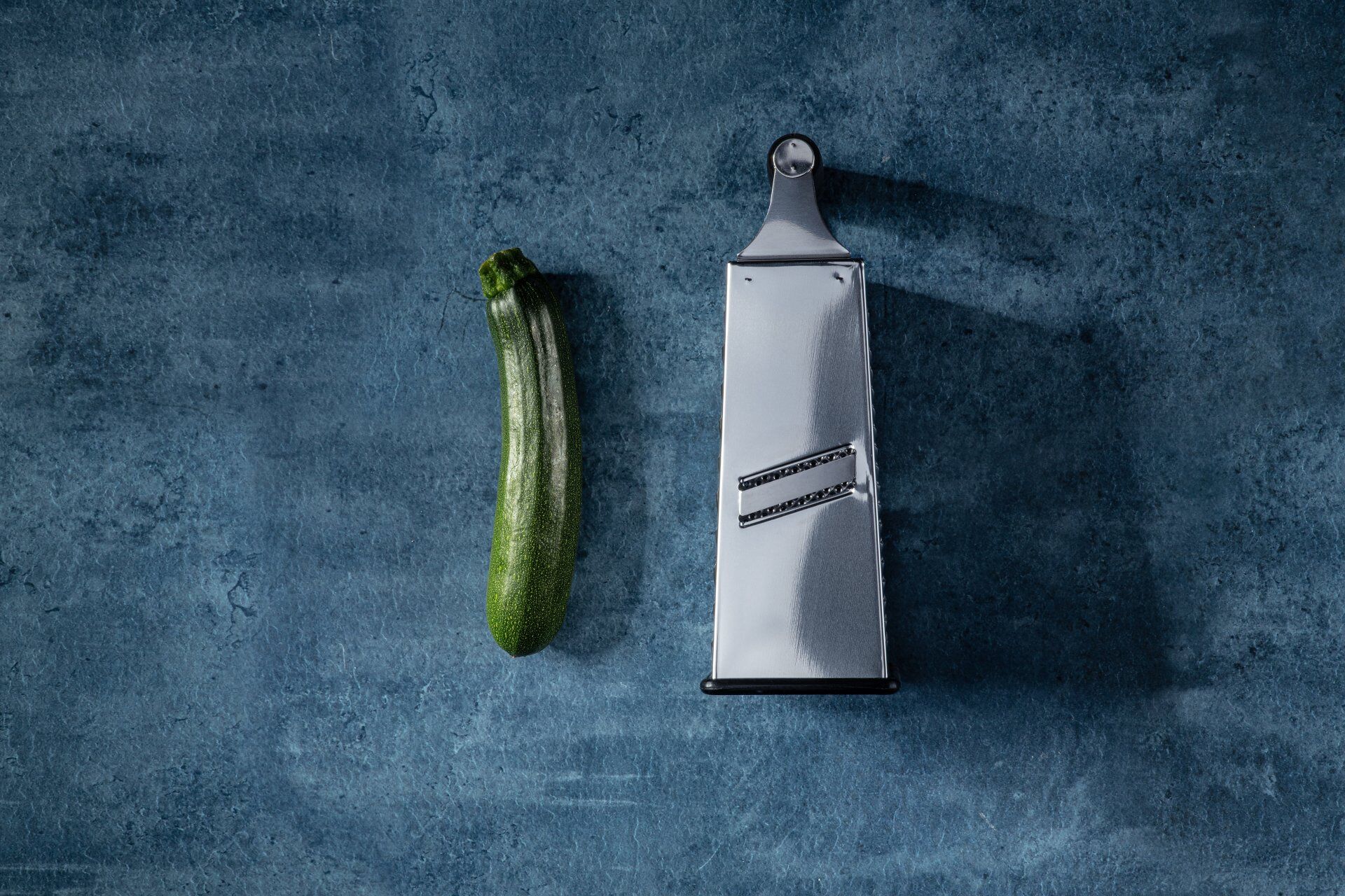 Ganze Zucchini neben einer Vierkantreibe auf blauer Fläche vor dem Zucchini einmachen Ganze Zucchini neben einer Vierkantreibe auf blauer Fläche vor dem Zucchini einmachen