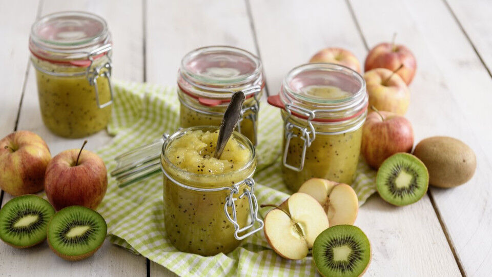 Apfelmus mit Kiwi in vier Einmachgläsern mit Bügelverschluss, daneben ganze und halbierte Äpfel und Kiwis