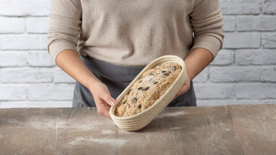Brotteig mit Kernen in einem länglichen Gärkörbchen, gehalten von einer Frau