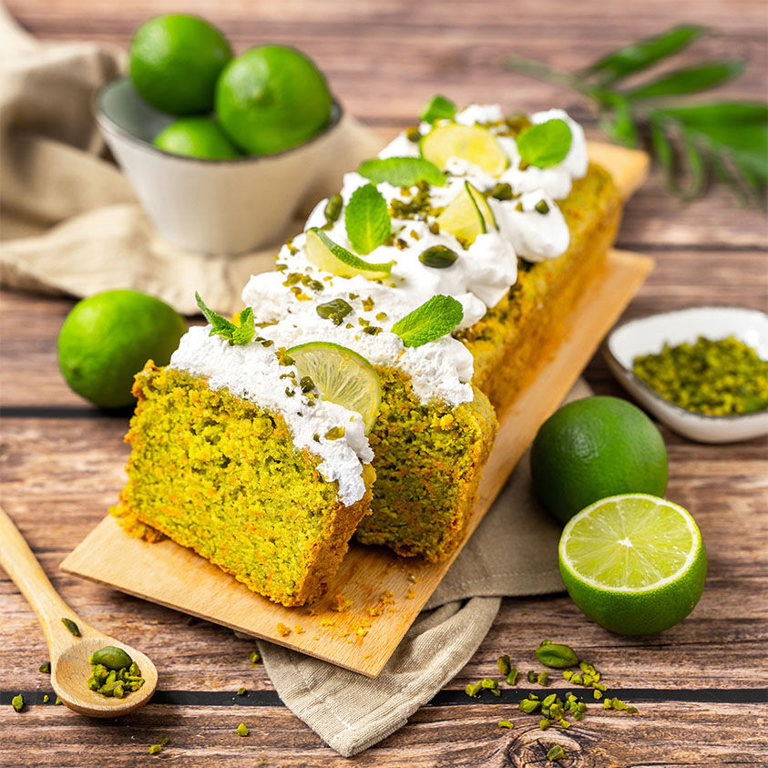Angeschnittener veganer Carrot Cake auf einem Holzbrett, daneben Limetten und Pistazien in Schälchen