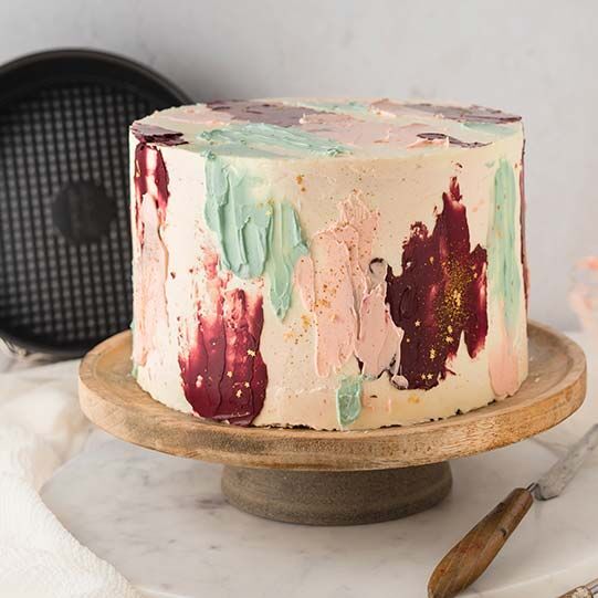 Bunte Flecken-Torte auf einer Tortenplatte aus Holz, daneben ein Dekorierspatel